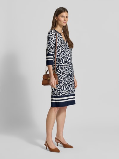Modelka kobieca nosi knielanges Tunikakleid mit V-Ausschnitt und 3/4-Arm od Christian Berg Woman Selection, mieszanka materiałów ze stretchem, prosty krój, długość do kolan, wzór allover, dekolt V, długość tyłu w rozmiarze 36: 92 cm, szerokość w rozmiarze 36: 41 cm, długość rękawa w rozmiarze 36: 42 cm, kolor granatowo-biały, widok całej sylwetki z przodu.