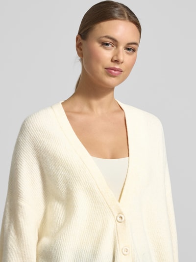 Frauenmodel trägt Relaxed Fit Cardigan mit Woll-Anteil Modell 'Damsville' von American Vintage, cremefarben, Strick-Optik mit V-Ausschnitt, elastischer Material-Mix, Knopfleiste, Nahaufnahme Oberkörper.