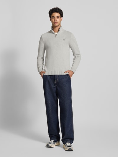 Męski model nosi Slim Fit Pullover in Strick-Optik von Superdry, szary sweter o kroju slim fit, stójka, zamek błyskawiczny, wyhaftowane logo, długość tyłu w rozmiarze M: 70 cm, długość rękawa w rozmiarze M: 65 cm, imitacja dzianiny, widok całej sylwetki z przodu.