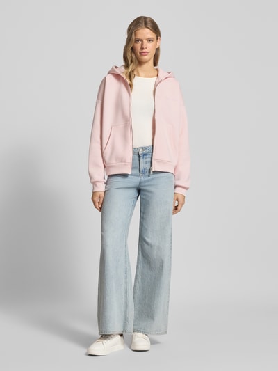 Frauenmodel trägt Oversized Sweatjacke mit Kapuze von Review, Material-Mix mit Baumwoll-Anteil, Label-Print, Eingrifftaschen, gerippte Abschlüsse, Reißverschluss, hellrosa, gerade Formen, glatte Textur, Ganzkörperansicht von vorne.