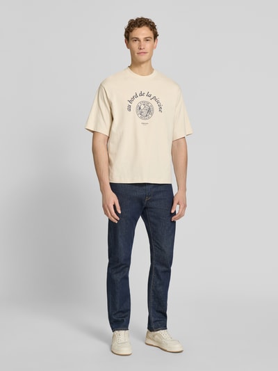Männermodel trägt Relaxed Fit T-Shirt aus reiner Baumwolle Modell 'BOXYNORMAN GRAPHIC' von SELECTED HOMME in Beige mit Motiv-Print, Ganzkörperansicht von vorne.