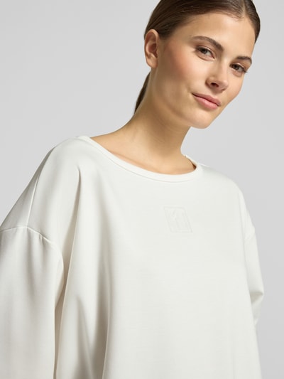Frauenmodel trägt T-Shirt mit Rundhalsausschnitt und Logo von monari, Damen T-Shirt im Relaxed-Fit aus Material-Mix mit Viskose-Anteil, Offwhite, glatte Textur, Nahaufnahme Oberkörper.