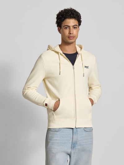 Männermodel trägt Regular Fit Sweatjacke mit Logo-Stitching von Superdry aus Baumwoll-Mix in Creme, mit Kapuze, Reißverschluss, Kängurutasche, elastische Abschlüsse, Herren, Ganzkörperansicht von vorne.