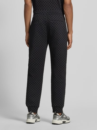 Männermodel trägt Sweatpants im Allover-Look mit Logo-Muster Modell 'PANTALONI' von Armani Exchange, Ganzkörperansicht Rückansicht. Baumwoll-Mix mit Stretch-Anteil, Regular Fit, elastischer Bund, Tunnelzug, dunkle Farbtöne, grafische Muster, strukturierte Textur.