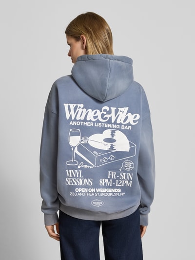 Kobiece model prezentuje Oversized Hoodie mit Känguru-Tasche Modell 'Wine & Vibe' od ANOTHER COTTON LAB, mieszanka bawełny, efekt znoszenia, nadruk z logo, kieszeń kangurka, widok całej sylwetki z tyłu.