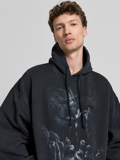 Männermodel trägt Oversized Hoodie aus Baumwoll-Mix von VETEMENTS, schwarz mit Motiv-Print, Destroyed-Look, Nahaufnahme Oberkörper.