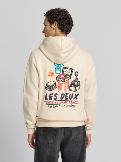 Männermodel trägt Hoodie aus reiner Baumwolle mit Logo-Print Modell 'Record' von Les Deux in Beige, farbiger Print, Ganzkörperansicht Rückansicht. Herren Hoodie von Les Deux, Regular Fit, Kapuze mit Tunnelzug, langärmlig, gerippte Abschlüsse, Kängurutasche, Logo-Label, Rückenlänge 70 cm, Ärmellänge 74 cm.