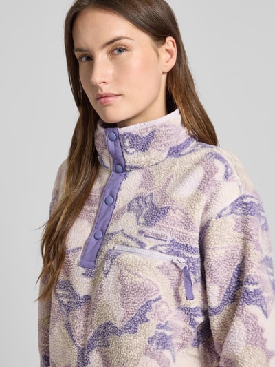 Modelka kobieca ma na sobie Regular Fit Pullover mit Logo-Patch Modell 'Helvetia' od Columbia w fasonie krój regular fit, wygląd polaru, elastyczne mankiety, kieszeń na piersi z zamkiem, listwa guzikowa, stójka, pastelowe odcienie fioletu i beżu, tekstura miękka. Zbliżenie na górną część ciała.
