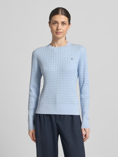 Kobiece modelka ma na sobie Slim Fit Pullover aus reiner Baumwolle von Tommy Hilfiger, efekt jednolity kolor, imitacja dzianiny, krój slim fit, wzór ściegu warkoczowego, detal z logo, okrągły dekolt, długi rękaw, prążkowane zakończenia, dzianina z bawełny; widok całej sylwetki z przodu.