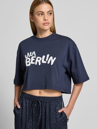 Frauenmodel trägt Cropped T-Shirt mit Logo-Print von Lala Berlin in Dunkelblau, Reine Baumwolle, Rundhalsausschnitt, Halbarm, Ganzkörperansicht von vorne.