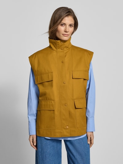 Modelka kobieca nosi Oversized Weste mit aufgesetzten Taschen - curated by Tinvcb od SIC, kolor musztardowy, stebnowane kieszenie z patkami, stójka, zapięcie na guziki, relaxed fit, długość tyłu w rozmiarze 36: 67 cm, szerokość ramion w rozmiarze 36: 52 cm, szerokość w biuście w rozmiarze 36: 59 cm, widok całej sylwetki z przodu.