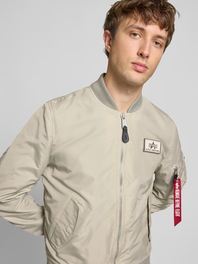 Męski model nosi Regular Fit Bomberjacke mit Logo-Patch von Alpha Industries w beżowym kolorze, wpuszczane kieszenie, zamek błyskawiczny, naszywka z logo, kieszeń na rękawie, stójka, krój regular fit, długość tyłu w rozmiarze M: 65 cm, szerokość ramion w rozmiarze M: 45 cm, szerokość klatki piersiowej w rozmiarze M: 54 cm, długość rękawów w rozmiarze M: 69 cm, całkowity widok przód.