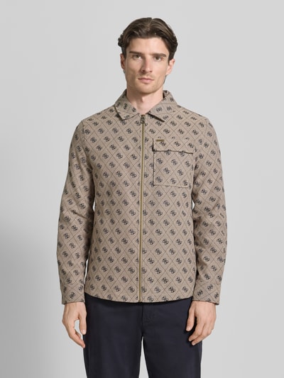 Männermodel trägt Regular Fit Jacke mit Brusttasche von Guess, beige mit schwarzem Allover-Logo-Stitching, Baumwoll-Mix, Reißverschluss, Eingrifftaschen, Umlegekragen, Ganzkörperansicht von vorne. Rückenlänge 73 cm, Schulterbreite 45 cm, Brustweite 51 cm, Ärmellänge 65 cm bei Größe M.