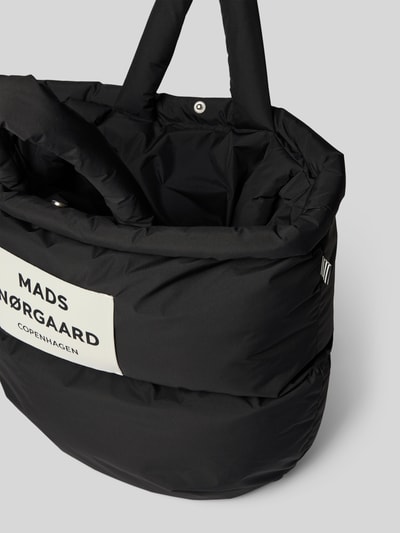 Produktfoto, Handtasche mit Label-Detail und Steppnähten von MADS NØRGAARD, schwarze glatte Oberfläche, sichtbarer Tragehenkel, Nahaufnahme. Breite 42 cm, Länge 32 cm, Tiefe 15 cm, Schulterriemen 66 cm.