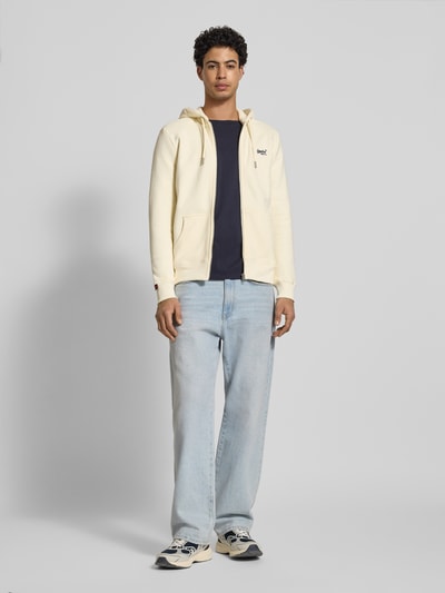 Männermodel trägt Regular Fit Sweatjacke mit Logo-Stitching von Superdry aus Baumwoll-Mix, Kapuze mit Tunnelzug, Reißverschluss, geteilte Kängurutasche, elastische Abschlüsse, helle Farben, glatte Textur, Rückenlänge 69 cm, Ärmellänge 65 cm, Ganzkörperansicht von vorne.