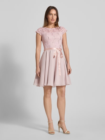 Kobiece modelka nosi Cocktailkleid mit Spitzenbesatz und Bindegürtel od SWING w kolorze pudrowego różu, materiał z dodatkiem stretchu, fason A-linia, minikleid, ozdobiony cekinami i koronką, na tle jednolitego szarego, całopostaciowy widok z przodu.