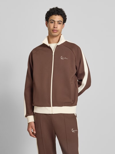 Männermodel trägt Sweatjacke mit Stehkragen von KARL KANI in Braun mit Material-Mix, Raglanärmel, Label-Stitching, Seitentaschen, Reißverschluss, lockere Passform, elastische Abschlüsse, Ganzkörperansicht von vorne.