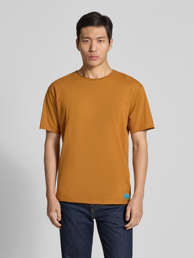 Männermodel trägt T-Shirt mit Rundhalsausschnitt und Label-Detail von Scotch & Soda in Braun, reine Baumwolle, Regular-Fit, Ganzkörperansicht von vorne.