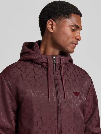 Männermodel trägt Regular Fit Jacke mit Allover-Logo-Print von Emporio Armani in Dunkelrot, Kapuze, Logo-Applikation, Nahaufnahme Oberkörper.