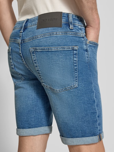 Männermodel trägt Regular Fit Jeansshorts aus Baumwoll-Mix Modell 'PLY' von Only & Sons in Denim-Optik mit Label-Patch, 5-Pocket-Design, Beinabschluss mit Umschlag, Detailaufnahme Rückansicht.