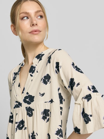 Frauenmodel trägt ausgestelltes Blusenkleid aus reiner Baumwolle Modell 'DICTHE' von Vero Moda mit Allover-Muster, helle Grundfarbe, schwarze florale Formen, Henley-Ausschnitt, 1/2-Trompetenärmel, leichte strukturierte Textur, Nahaufnahme Oberkörper.