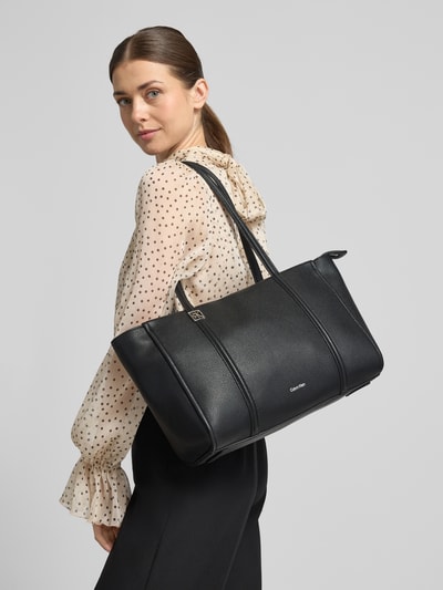 Kobieta modelka nosi Shopper mit Label-Detail von CK Calvin Klein w kolorze czarnym, faktura skóry, szerokość w rozmiarze 47 cm, długość 26 cm, głębokość 16 cm, długość paska na ramię 63 cm, główna komora na zamek, wewnętrzna kieszeń na zamek, uchwyty, widok całej sylwetki z tyłu.