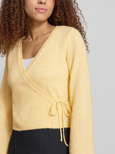 Frauenmodel trägt Regular Fit Strickjacke mit Woll-Anteil Modell 'COLO' von Only in Melange-Optik, Wickeloptik, Bindeband, V-Ausschnitt, Langarm, gerippter Ärmelabschluss und Rumpfabschluss, gestrickte Materialverarbeitung, Nahaufnahme Oberkörper.