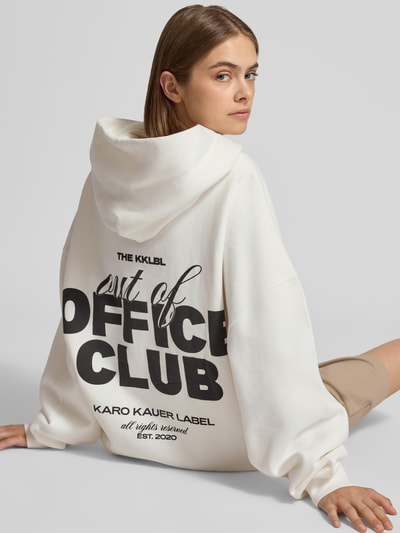 Frauenmodel trägt Oversized Hoodie mit Känguru-Tasche von Karo Kauer, cremefarben, Label-Print, strukturierte Baumwolle, Ganzkörperansicht Rückansicht.