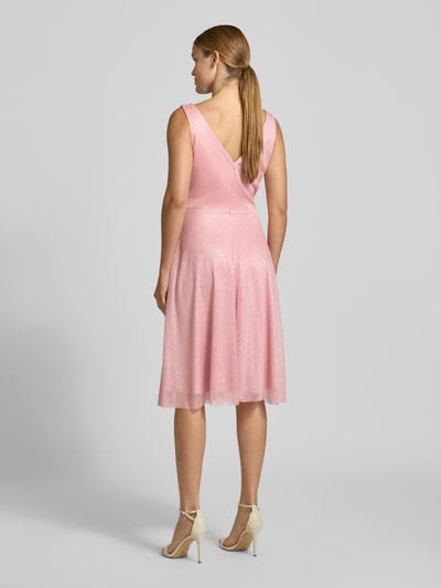 Frauenmodel trägt Knielanges Cocktailkleid mit V-Ausschnitt von TROYDEN COLLECTION in Rosa, Chiffon, tailliert, knielang, breite Träger, Rückseitiger V-Ausschnitt, Reißverschluss, feine Textur, dezenter Glanz, Ganzkörperansicht Rückansicht.