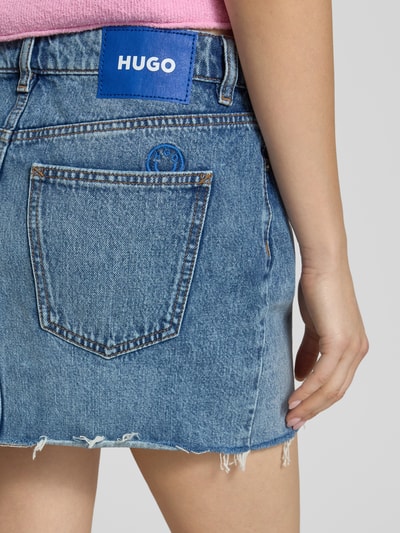 Frauenmodel trägt Mini-Jeansrock aus reiner Baumwolle Modell 'GALANIE_1_B' von Hugo Blue in Denim-Optik mit Nieten, Label-Patch, Logo-Stitching, Gürtelschlaufen, Kontrastnaht, Knopfleiste, offener Kante und 5-Pocket-Design, Ganzkörperansicht Rückansicht.