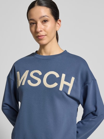 Vrouwenmodel draagt Sweatshirt met oversneden schouders van MSCH Copenhagen in blauw, katoenmix, rechte fit, model 'Mela Briena', ronde hals, borstlogo. Close-up bovenlichaam.