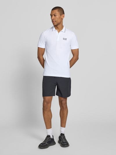 Männermodel trägt Regular Fit Poloshirt mit Logo-Print von EA7 Emporio Armani in Weiß, Kurzarm, schwarzer Shorts, Ganzkörperansicht von vorne.
