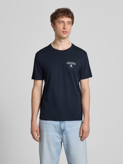 Männermodel trägt T-Shirt mit Logo und Rundhalsausschnitt von CHAMPION in Dunkelblau, Reine Baumwolle, Regular-Fit, Ganzkörperansicht von vorne. Rückenlänge bei Größe M: 61 cm.