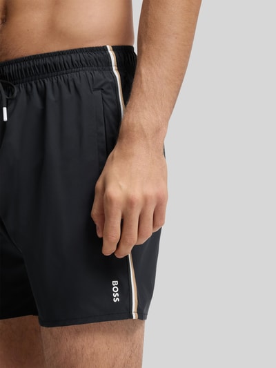 Männermodel trägt Badeshorts mit Tunnelzug Modell 'ICONIC' von BOSS, unifarben, Kontraststreifen, Label Print, Nahaufnahme Oberschenkel.