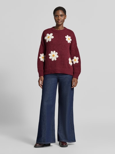 Kobieta prezentuje sweter z dzianiny 'Oversized Strickpullover mit floralen Stitchings' od Apricot, bordowy, kwiatowe hafty, okrągły dekolt, krój oversized, prążkowane wykończenia, długość tyłu 62 cm, długość rękawów 43 cm, widok całej sylwetki z przodu.