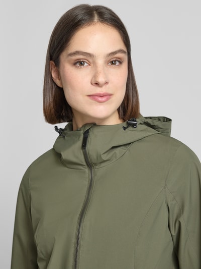 Frauenmodel trägt Regular Fit Regenmantel mit Logo-Detail von G.I.G.A. DX in Olivgrün, glatte Oberfläche, Nahaufnahme Oberkörper. Rückenlänge 76 cm, Schulterbreite 40 cm, Brustweite 50 cm, Ärmellänge 66 cm, Kapuze mit Tunnelzug, Zweiwege-Reißverschluss, Eingrifftaschen mit Reißverschluss.