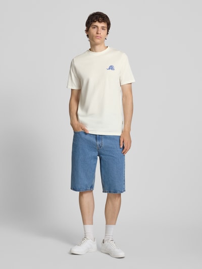 Männermodel trägt T-Shirt mit Motiv-Print Modell 'SUN ISLAND' von Blend in Creme, kombiniert mit blauen Denim-Shorts, Ganzkörperansicht von vorne. Herren T-Shirt aus reiner Baumwolle im Regular Fit mit Rundhalsausschnitt, Motiv-Print auf der Brust, Rückenlänge bei Größe M 71 cm.