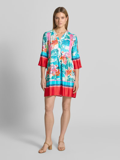 Frauenmodel trägt Relaxed Fit Minikleid aus reiner Viskose Modell 'Breeze' von miss goodlife mit floraler Allover-Print, Halbarm, V-Ausschnitt, Knopfleiste und Falten-Optik in leuchtendem Türkis, Rot und Pink, Ganzkörperansicht von vorne. Rückenlänge bei Größe S: 40 cm, Brustweite: 39 cm, Ärmellänge: 46 cm.