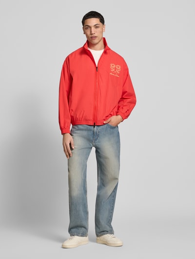 Męski model nosi Segeljacke mit Stehkragen und Logo-Stitching von ANOTHER COTTON LAB, bawełna, prosty krój, czerwona, zamek błyskawiczny dwustronny, wyhaftowane logo, elastyczne wykończenia, długość tyłu w rozmiarze M 63 cm, szerokość ramion w rozmiarze M 60 cm, szerokość w rozmiarze M 63 cm, długość rękawów w rozmiarze M 55 cm, widok całej sylwetki z przodu.
