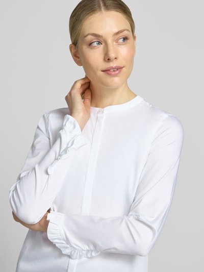 Modelka nosi bluzkę z mieszanki bawełny z dodatkiem streczu MOS MOSH, Model 'Mattie', stójka, listwa guzikowa kryta, rękawy z falbanami, długość tyłu w rozmiarze S: 63 cm, długość rękawów w rozmiarze S: 62 cm, kolor biały, prosty krój, zbliżenie na górną część ciała z przodu.