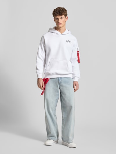 Männermodel trägt Hoodie mit Label-Details von Alpha Industries aus Baumwoll-Mix, lockerer Schnitt, Weiß, Känguru-Tasche, Ganzkörperansicht von vorne.
