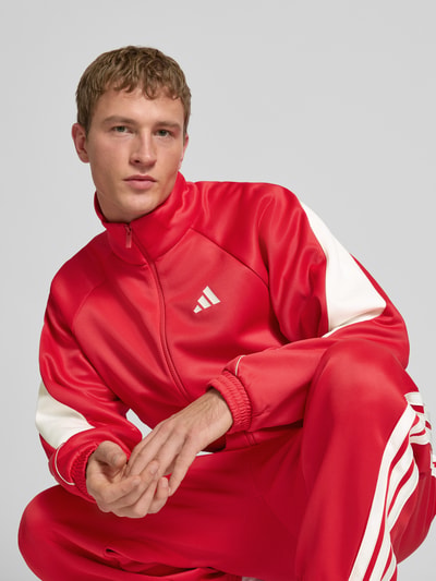 Männermodel trägt Trainingsjacke mit Stehkragen und Logo von adidas Sportswear in Rot mit weißen Details, Material-Mix, Nahaufnahme Oberkörper.