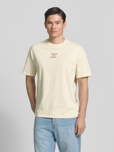 Męski model nosi T-Shirt z nadrukiem labelowym model 'GERGEI' od Samsøe Samsøe, bawełna, krój relaxed fit, jasny beż, okrągły dekolt, długość tyłu w rozm. M: 71 cm, widok całej sylwetki z przodu.