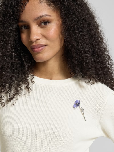 Frauenmodel trägt Slim Fit Strickpullover aus reiner Baumwolle Modell 'SABANNA' von HUGO in Creme mit floraler Brosche, Nahaufnahme Oberkörper.