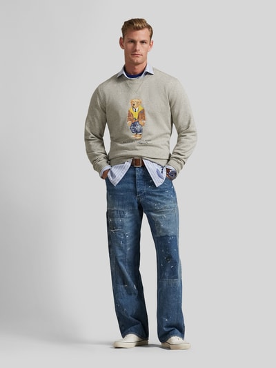 Męski model nosi Regular Fit Sweatshirt aus Baumwoll-Mix mit Polo Bear od Polo Ralph Lauren, melanż szary, print Polo-Bear, okrągły dekolt, długie rękawy i ściągacze, materiał sweat bawełniany, krój regular fit, widok całej sylwetki z przodu.