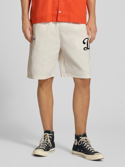 Männermodel trägt Relaxed Fit Shorts aus reiner Baumwolle von Champion in Beige mit elastischem Bund, Logo-Stitching, Nahaufnahme Unterkörper.