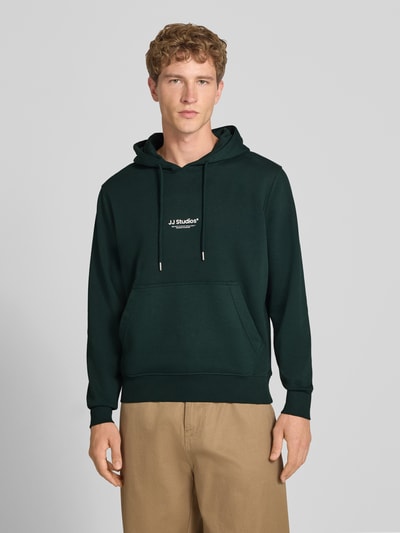 Männermodel trägt Regular Fit Hoodie aus Baumwoll-Mix Modell 'Soho' von JACK & JONES in Dunkelgrün mit Känguru-Tasche, Ganzkörperansicht von vorne. Baumwoll-Mix, Kapuze mit Tunnelzug, elastische Abschlüsse, Rückenlänge 66 cm, Ärmellänge 65 cm.