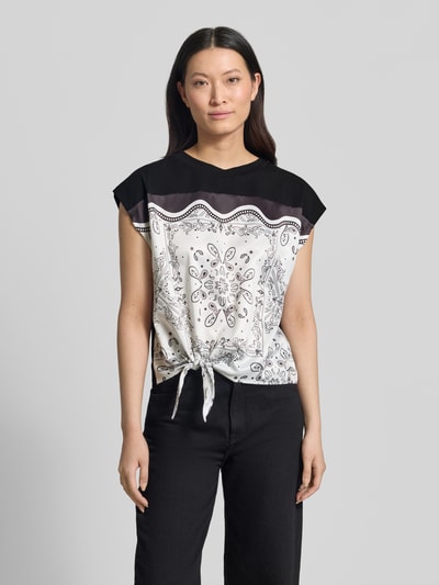 Frauenmodel trägt Blusenshirt mit Knotendetail von Christian Berg Woman Selection aus Satin, Regular-Fit, Rundhalsausschnitt, Rückenlänge 52 cm bei Größe 36, schwarz-weiß gemustert, Ganzkörperansicht von vorne.