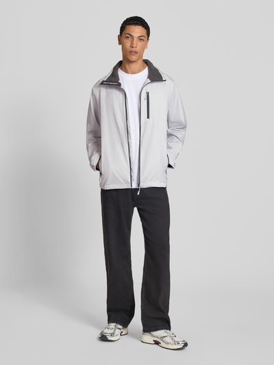 Männermodel trägt Regular Fit Jacke mit Stehkragen Modell 'Crew' von Helly Hansen in Grau, glatte Struktur, Ganzkörperansicht von vorne. Herren Jacke, Logo-Print, Napoleontasche, Eingrifftaschen mit Reißverschluss, Rückenlänge 73 cm, Schulterbreite 48 cm, Brustweite 56 cm, Ärmellänge 68 cm.