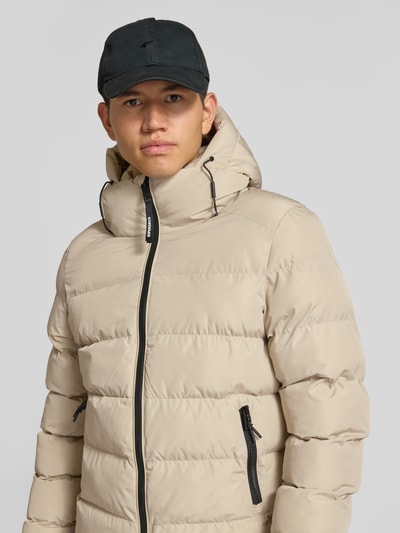 Mannenmodel draagt gewatteerd jack met labelstitching en capuchon van Superdry in beige met zwarte ritssluiting en steekzakken, recht model, detailopname bovenlichaam. Ruglengte maat M 67 cm, schouderbreedte 48 cm, borstwijdte 58 cm, mouwlengte 72 cm.